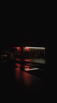 A dimly lit tattoo parlor with a red neon sign is reflected on the wet pavement. The surrounding area is dark, highlighting the glowing signage and creating a moody, atmospheric scene.