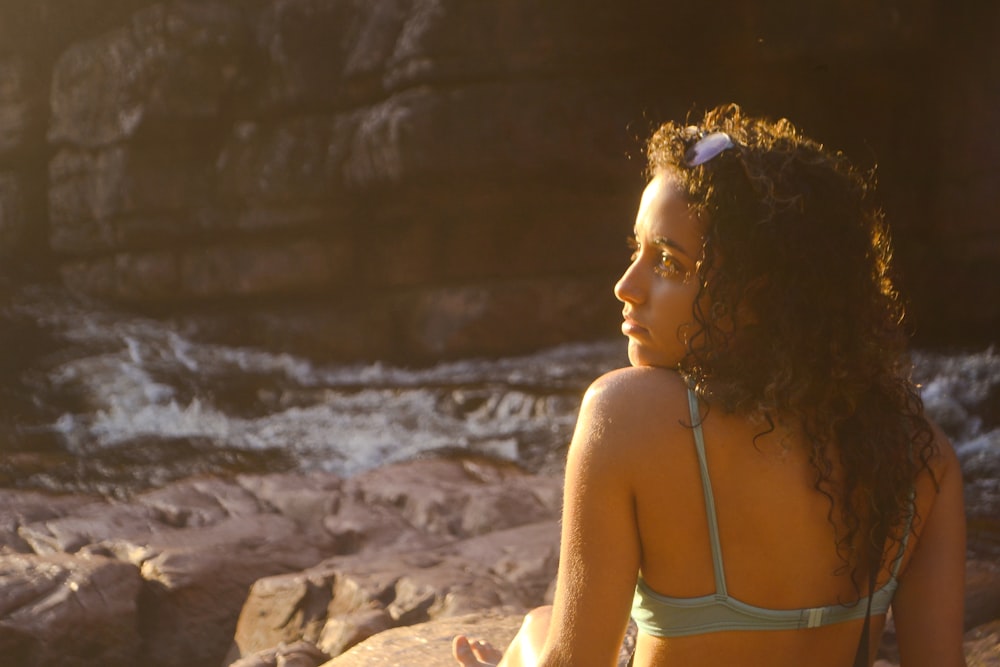 a woman in a bikini sitting on a rock next to a body of water