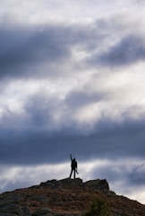 A person stands triumphantly on a rocky hilltop under a dramatic, cloudy sky. The individual is silhouetted against the light, raising one hand, suggesting a sense of accomplishment.