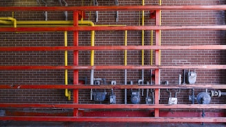 A setup of interconnected pipes and meters mounted on a brick wall, with a series of horizontal red beams. Vertical yellow pipes contrast against the red structure and the dark brick background, housing several utility meters and valves.