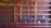 A setup of interconnected pipes and meters mounted on a brick wall, with a series of horizontal red beams. Vertical yellow pipes contrast against the red structure and the dark brick background, housing several utility meters and valves.