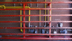 A setup of interconnected pipes and meters mounted on a brick wall, with a series of horizontal red beams. Vertical yellow pipes contrast against the red structure and the dark brick background, housing several utility meters and valves.