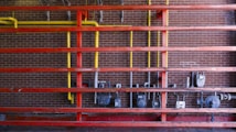 A setup of interconnected pipes and meters mounted on a brick wall, with a series of horizontal red beams. Vertical yellow pipes contrast against the red structure and the dark brick background, housing several utility meters and valves.