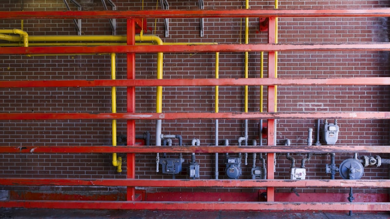A setup of interconnected pipes and meters mounted on a brick wall, with a series of horizontal red beams. Vertical yellow pipes contrast against the red structure and the dark brick background, housing several utility meters and valves.