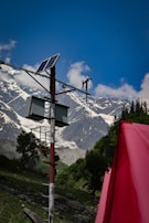 A solar panel is mounted on a pole with additional equipment, set against a backdrop of towering, snow-capped mountains. The sky is clear and blue, with a few scattered clouds. In the foreground, a bright red object, possibly a tent, adds contrast to the lush green landscape.