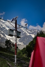 A solar panel is mounted on a pole with additional equipment, set against a backdrop of towering, snow-capped mountains. The sky is clear and blue, with a few scattered clouds. In the foreground, a bright red object, possibly a tent, adds contrast to the lush green landscape.