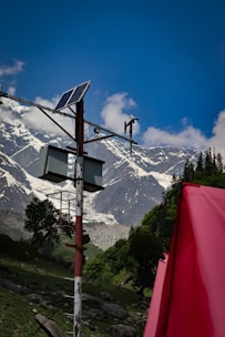 A solar panel is mounted on a pole with additional equipment, set against a backdrop of towering, snow-capped mountains. The sky is clear and blue, with a few scattered clouds. In the foreground, a bright red object, possibly a tent, adds contrast to the lush green landscape.
