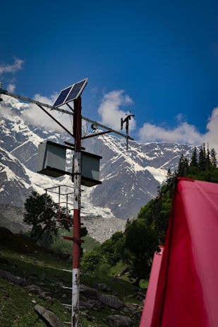 A solar panel is mounted on a pole with additional equipment, set against a backdrop of towering, snow-capped mountains. The sky is clear and blue, with a few scattered clouds. In the foreground, a bright red object, possibly a tent, adds contrast to the lush green landscape.
