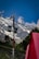 A solar panel is mounted on a pole with additional equipment, set against a backdrop of towering, snow-capped mountains. The sky is clear and blue, with a few scattered clouds. In the foreground, a bright red object, possibly a tent, adds contrast to the lush green landscape.