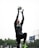A person is leaping into the air, reaching to catch a soccer ball with gloved hands. They are dressed in sportswear, including a black shirt with logos and black pants. The background shows a cloudy sky and blurred trees.