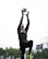 A person is leaping into the air, reaching to catch a soccer ball with gloved hands. They are dressed in sportswear, including a black shirt with logos and black pants. The background shows a cloudy sky and blurred trees.