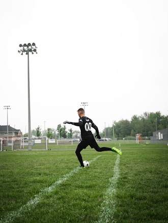 A person wearing a black sports outfit and neon green soccer shoes is playing soccer on a grass field. They are about to kick a white and black soccer ball. The field is empty with several floodlights and a building visible in the background. The sky is overcast and the atmosphere seems calm.