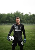 A soccer player is standing on a grassy field wearing a black goalkeeper uniform. He is holding a soccer ball in one hand and has a serious expression. The background features dense, green trees under an overcast sky.