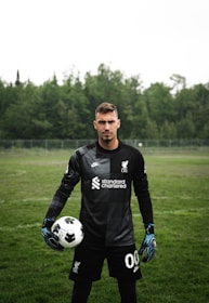 A soccer player is standing on a grassy field wearing a black goalkeeper uniform. He is holding a soccer ball in one hand and has a serious expression. The background features dense, green trees under an overcast sky.