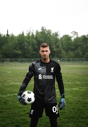 A soccer player is standing on a grassy field wearing a black goalkeeper uniform. He is holding a soccer ball in one hand and has a serious expression. The background features dense, green trees under an overcast sky.