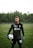 A soccer player is standing on a grassy field wearing a black goalkeeper uniform. He is holding a soccer ball in one hand and has a serious expression. The background features dense, green trees under an overcast sky.