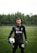 A soccer player is standing on a grassy field wearing a black goalkeeper uniform. He is holding a soccer ball in one hand and has a serious expression. The background features dense, green trees under an overcast sky.