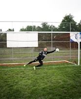 A soccer goalie dressed in black athletic gear is leaping horizontally, attempting to catch a soccer ball in front of a goal. The net is surrounded by metal fencing and the ground is covered with green grass. Trees and a cloudy sky form the backdrop.