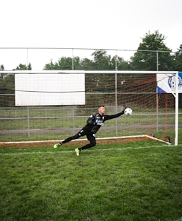 A soccer goalie dressed in black athletic gear is leaping horizontally, attempting to catch a soccer ball in front of a goal. The net is surrounded by metal fencing and the ground is covered with green grass. Trees and a cloudy sky form the backdrop.