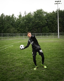 A soccer player is on a grassy field, wearing a black sports outfit and gloves, and is holding a black-and-white soccer ball. The background features a tall fence and a line of dense, green trees under a cloudy sky.