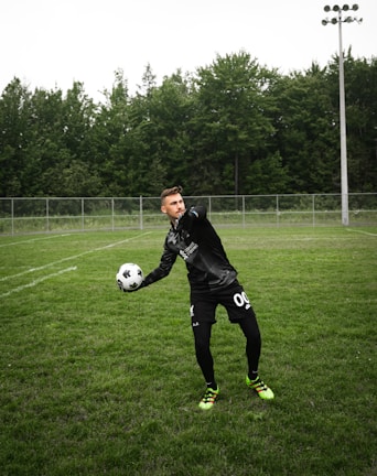 A soccer player is on a grassy field, wearing a black sports outfit and gloves, and is holding a black-and-white soccer ball. The background features a tall fence and a line of dense, green trees under a cloudy sky.