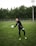 A soccer player is on a grassy field, wearing a black sports outfit and gloves, and is holding a black-and-white soccer ball. The background features a tall fence and a line of dense, green trees under a cloudy sky.