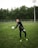 A soccer player is on a grassy field, wearing a black sports outfit and gloves, and is holding a black-and-white soccer ball. The background features a tall fence and a line of dense, green trees under a cloudy sky.