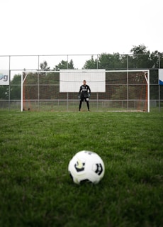 A soccer ball is positioned in the foreground on a grassy field, with a goalkeeper standing in front of the goal in the background. The field is surrounded by a tall fence and trees are visible beyond the fence.