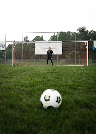 A soccer ball is positioned in the foreground on a grassy field, with a goalkeeper standing in front of the goal in the background. The field is surrounded by a tall fence and trees are visible beyond the fence.