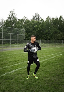 A soccer player in a black goalkeeper uniform stands on a grassy field, holding a soccer ball with both hands. A chain-link fence and dense green trees are visible in the background.