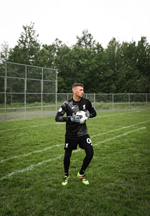 A soccer player in a black goalkeeper uniform stands on a grassy field, holding a soccer ball with both hands. A chain-link fence and dense green trees are visible in the background.