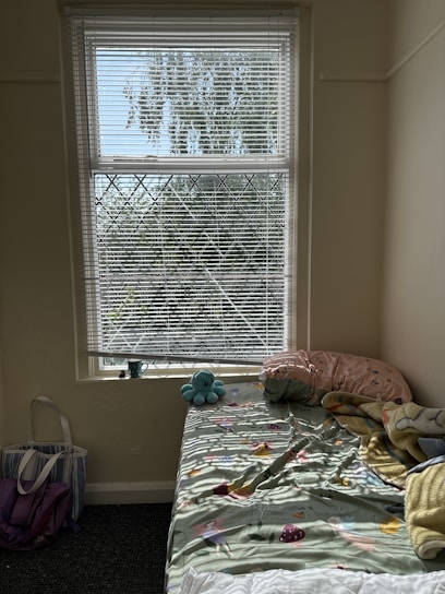 A cozy bedroom corner featuring a bed with colorful bedding, a stuffed toy placed on top. A window with blinds allows natural light to filter in, creating patterns on the bed. A tote bag and some objects rest on the floor nearby.