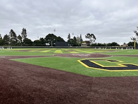 A baseball field with artificial turf infield and outfield, featuring a large yellow 'C' symbol in a corner. There are people, likely players, spread out along the outfield and another group near the left edge. Tall trees and some structures, including floodlights and a scoreboard, are visible in the background under a cloudy sky.