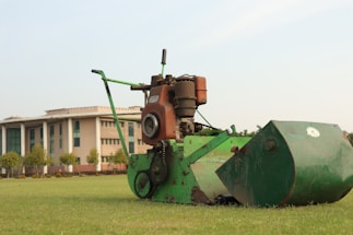 A green lawn mower with a rust-colored engine is positioned on a neatly mowed grass field. In the background, there is a large building with a modern architectural style, featuring tall pillars and numerous windows. Trees line the area near the building.