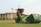 A green lawn mower with a rust-colored engine is positioned on a neatly mowed grass field. In the background, there is a large building with a modern architectural style, featuring tall pillars and numerous windows. Trees line the area near the building.