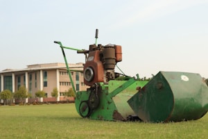 A green lawn mower with a rust-colored engine is positioned on a neatly mowed grass field. In the background, there is a large building with a modern architectural style, featuring tall pillars and numerous windows. Trees line the area near the building.