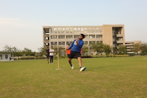A person is playing cricket on a well-maintained grassy field. The action captured shows a bowling motion, with the player mid-delivery. In the background, a large, multi-story building is visible. There are a few other people present, observing the game.
