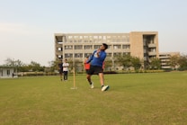 A person is playing cricket on a well-maintained grassy field. The action captured shows a bowling motion, with the player mid-delivery. In the background, a large, multi-story building is visible. There are a few other people present, observing the game.