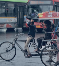 A street scene in an urban area with a bicycle rickshaw carrying three people, including a child. The background includes two large buses with vibrant colors. It appears to be raining lightly, as evidenced by the wet road and the people covering themselves.