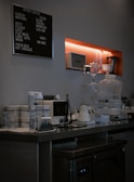 A modern coffee bar setup with various equipment including a coffee machine, kettle, and glass jars. A menu board on the wall lists different coffee options such as espresso, americano, and mocha. Shelving holds additional items like a speaker and cups.