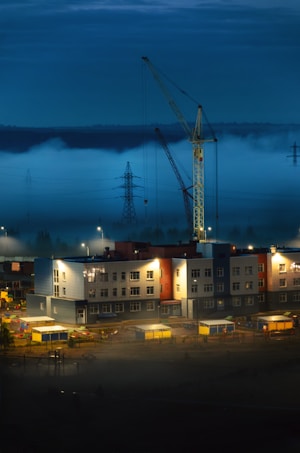 A construction site is illuminated under a dusky sky with a crane towering over a modern building. Mist rolls across the landscape, creating an ethereal atmosphere. The structure is surrounded by scattered construction materials and equipment, while power lines stretch into the horizon.