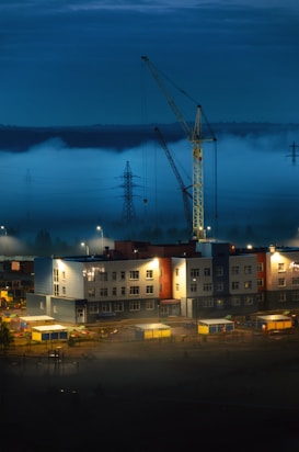 A construction site is illuminated under a dusky sky with a crane towering over a modern building. Mist rolls across the landscape, creating an ethereal atmosphere. The structure is surrounded by scattered construction materials and equipment, while power lines stretch into the horizon.