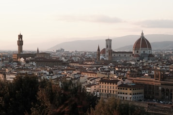 A panoramic view of an Italian city featuring prominent historical architecture, including a large cathedral with a domed roof and a tall bell tower. The buildings are closely packed, showcasing red-tiled roofs and stone structures with hills and cloudy skies in the background.