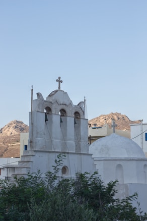 A whitewashed church structure with a bell tower featuring three arches, adorned with a cross on top. The church is surrounded by greenery with a mountainous backdrop under a clear blue sky.