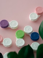 Several contact lens cases in various colors such as purple, green, blue, and pink are laid out on a pale pink background. The cases are labeled with 'L' and 'R' for the left and right eye. There are also green leaves partially visible at the bottom.