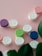 Several contact lens cases in various colors such as purple, green, blue, and pink are laid out on a pale pink background. The cases are labeled with 'L' and 'R' for the left and right eye. There are also green leaves partially visible at the bottom.