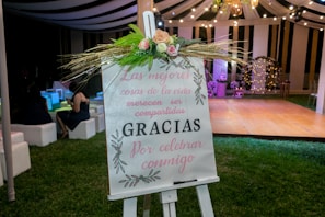 A decorative easel with a floral arrangement is placed on a grassy area at an event. The sign on the easel bears a message in Spanish, with elegant calligraphy and floral designs. In the background, a tent with draped fabric and string lights creates a festive atmosphere, and several people are seated on white cushioned seats, adding to the celebratory setting.