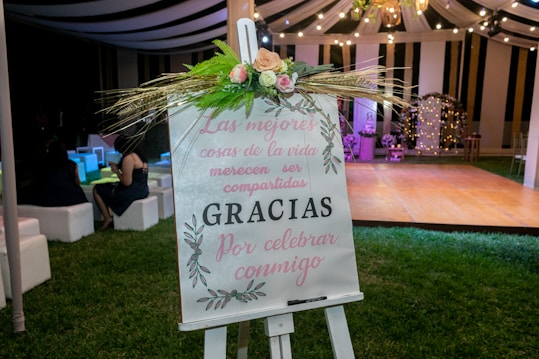 A decorative easel with a floral arrangement is placed on a grassy area at an event. The sign on the easel bears a message in Spanish, with elegant calligraphy and floral designs. In the background, a tent with draped fabric and string lights creates a festive atmosphere, and several people are seated on white cushioned seats, adding to the celebratory setting.