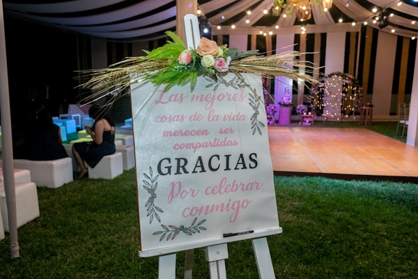 A decorative easel with a floral arrangement is placed on a grassy area at an event. The sign on the easel bears a message in Spanish, with elegant calligraphy and floral designs. In the background, a tent with draped fabric and string lights creates a festive atmosphere, and several people are seated on white cushioned seats, adding to the celebratory setting.