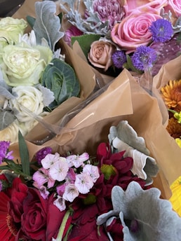 A collection of floral arrangements featuring a variety of flowers. Prominent colors include pink, purple, red, and white roses and carnations, along with green leaves and silver-grey dusty miller foliage. The bouquets are wrapped in brown paper.